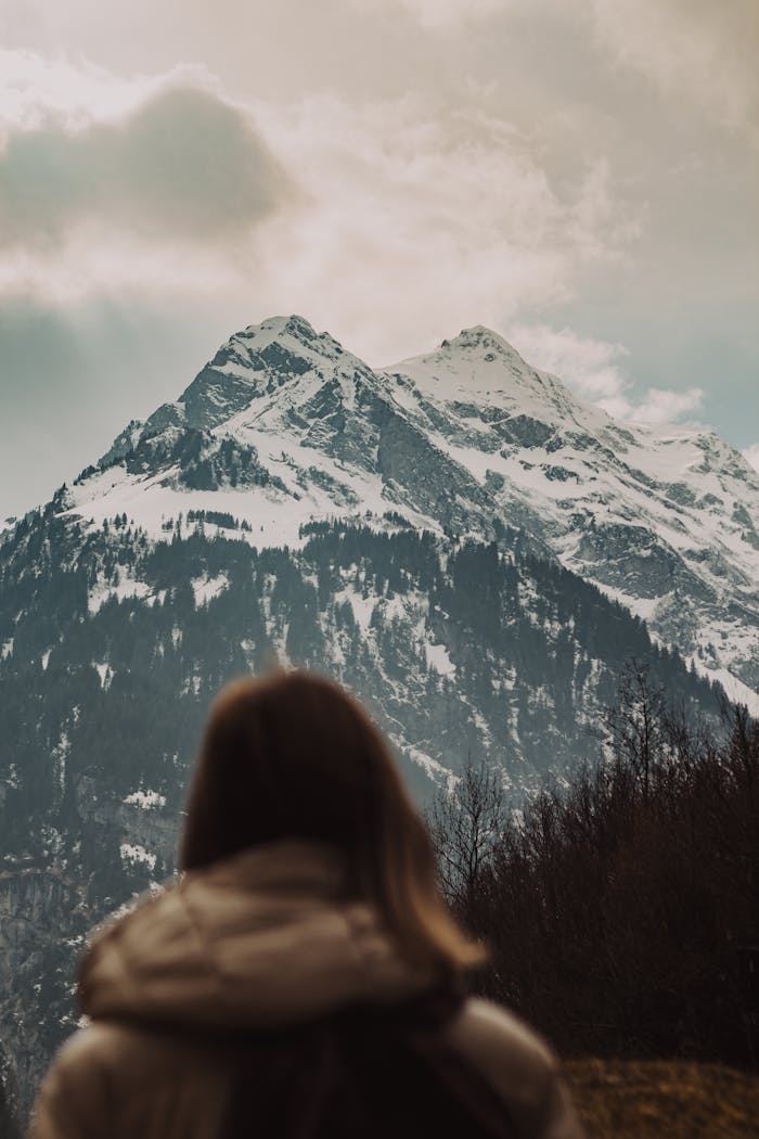 A woman in a coat looks at a majestic snow-capped mountain under cloudy skies.
