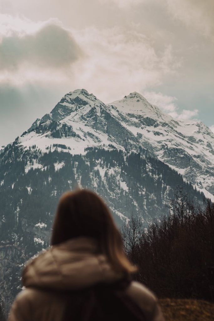 A woman in a coat looks at a majestic snow-capped mountain under cloudy skies.